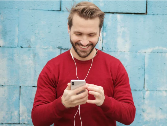 Man smiling while checking his smartphone, standing against a painted blue brick wall