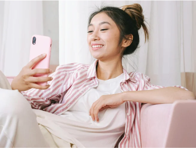 Woman relaxing on a sofa, smiling while taking a selfie with a pink smartphone